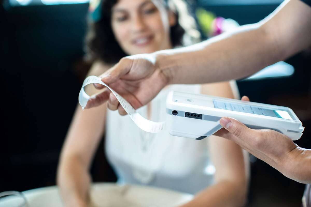 Una mano sostiene un terminal de pago digital y un recibo, con una persona borrosa sonriendo al fondo