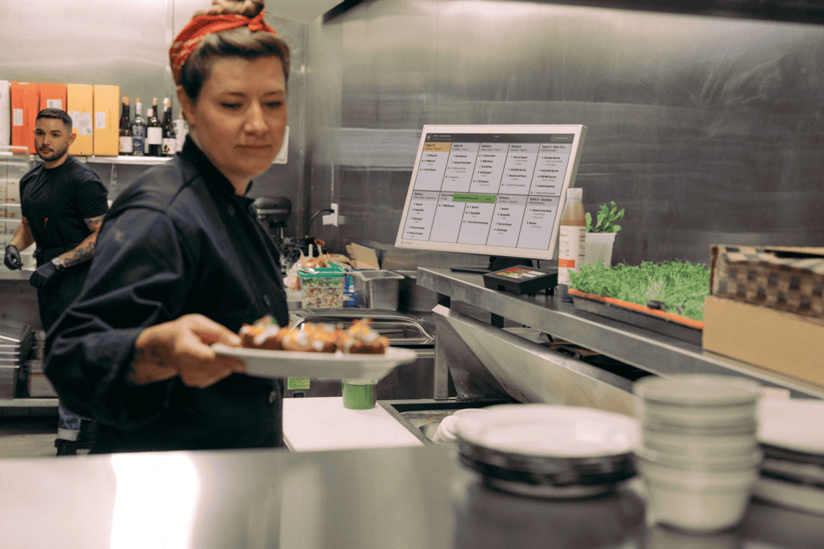 En una cocina profesional, un chef sostiene un plato de comida, mientras que al fondo se ve a otra persona y una pantalla digital para realizar pedidos