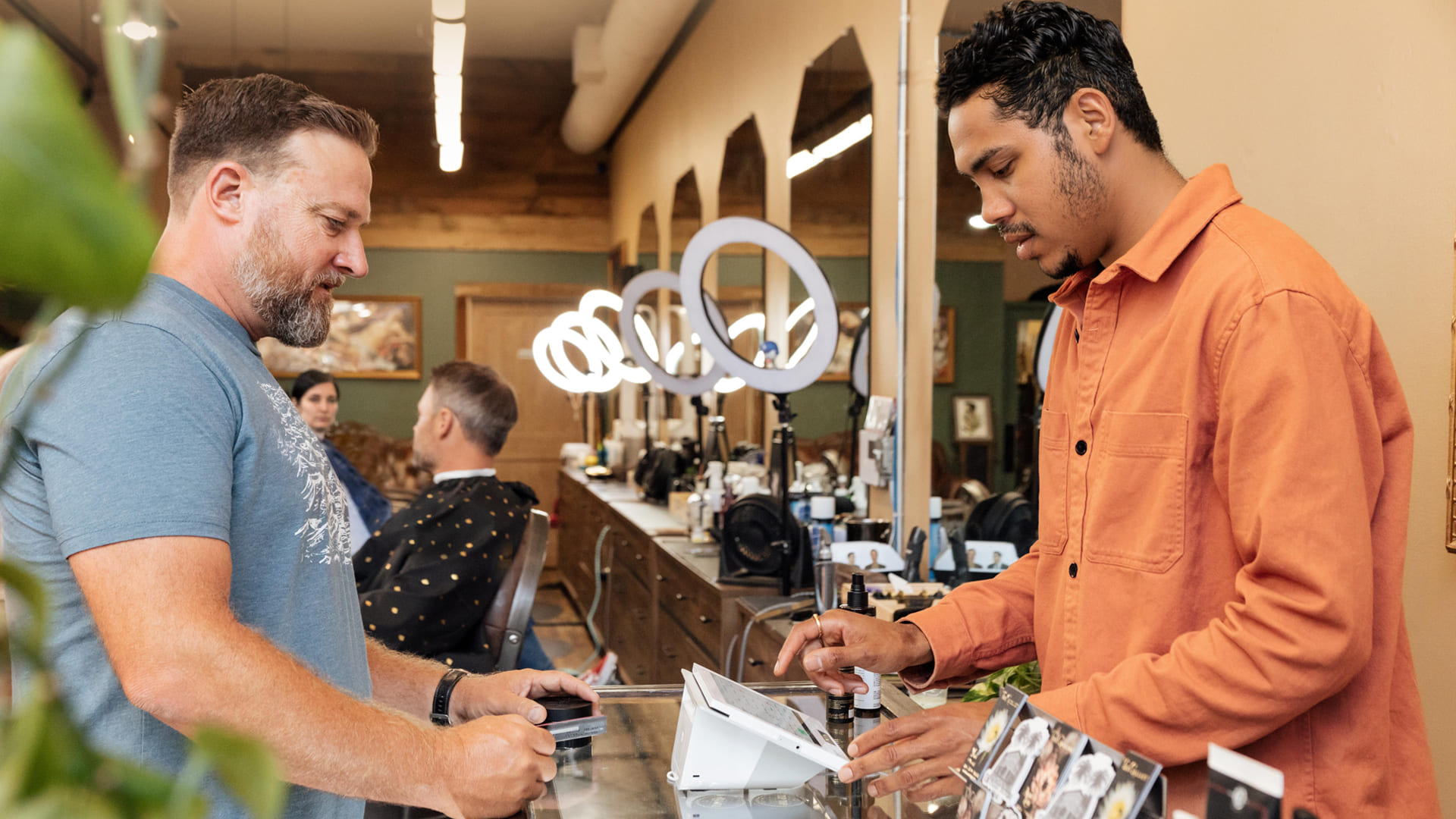 Cliente realizando un pago en la recepción de una barbería moderna mientras un empleado utiliza un sistema Clover POS con pantalla táctil, con luces circulares y sillones al fondo