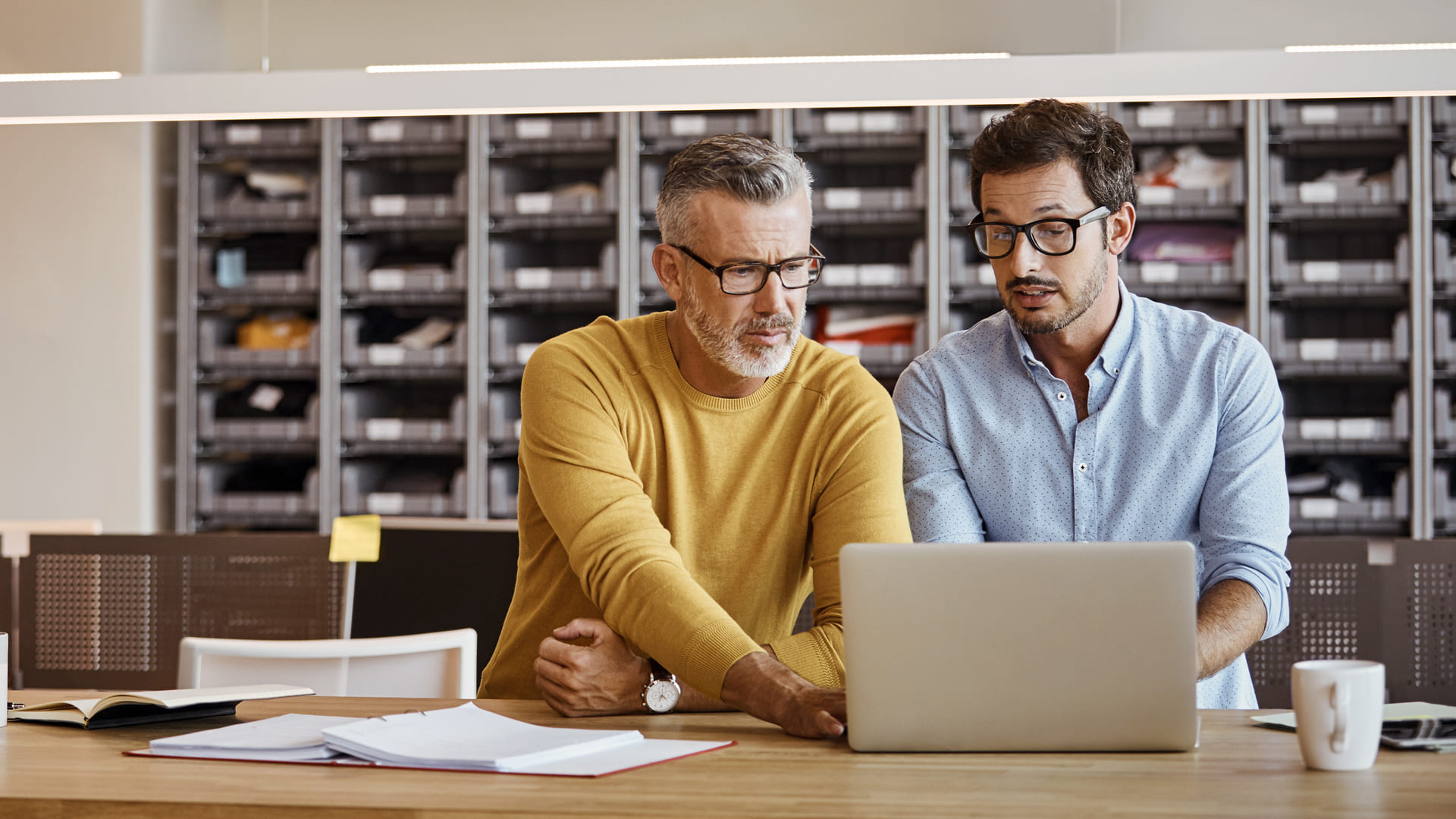 Dos hombres con gafas trabajan juntos en un escritorio, mirando la pantalla de un ordenador portátil en una oficina moderna con estanterías al fondo.