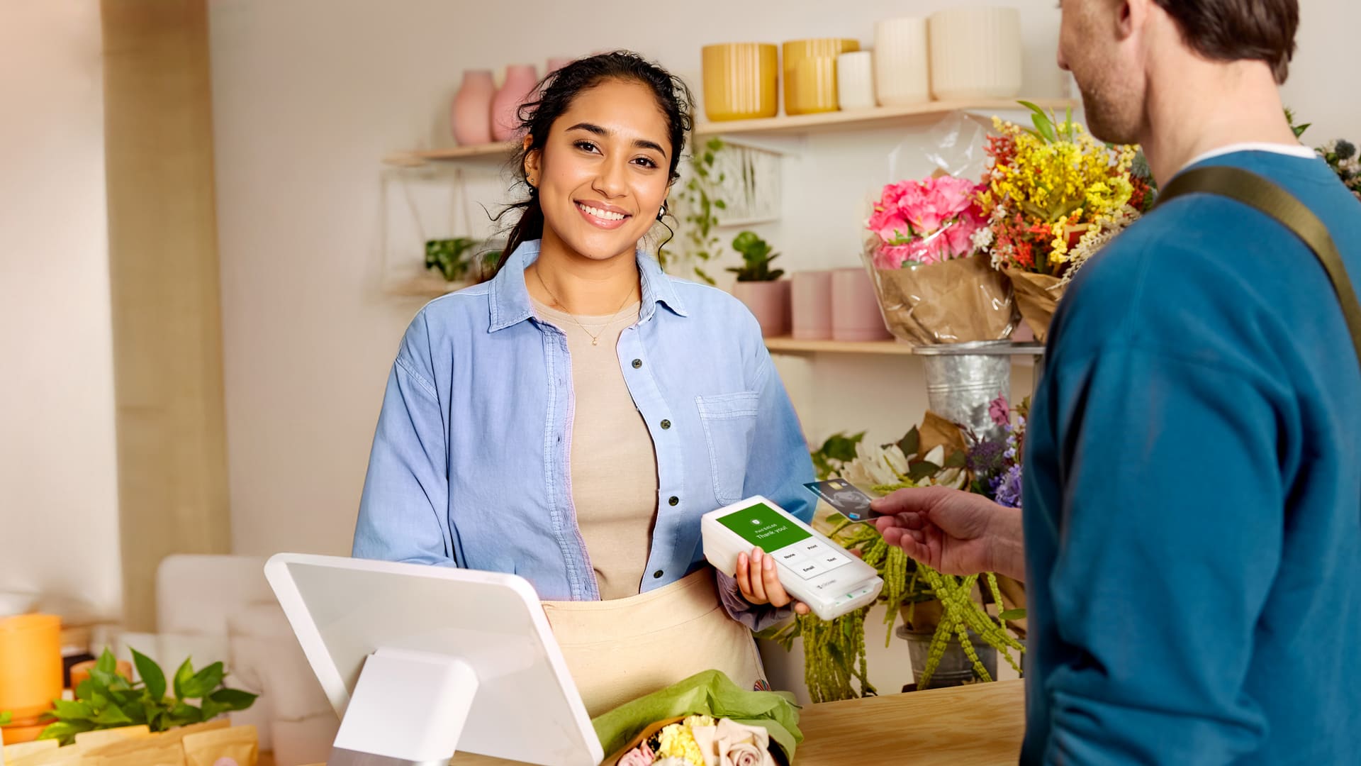 Florista sosteniendo un terminal portátil de pago Clover mientras un cliente acerca una tarjeta de crédito en una floristería llena de ramos coloridos y plantas