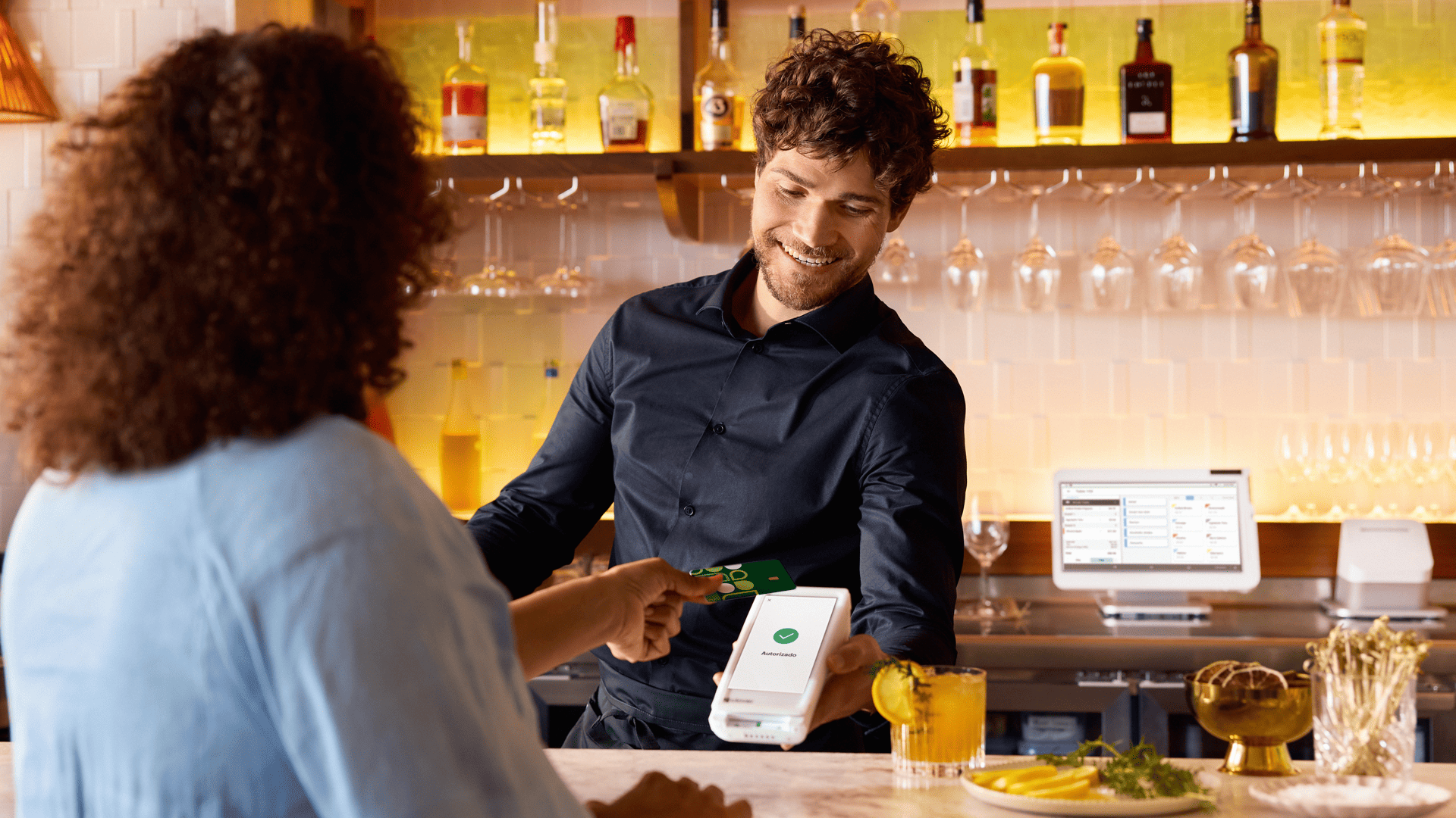 Barman procesando pago con tarjeta en un bar moderno con dispositivo POS, cliente entregando tarjeta de crédito.