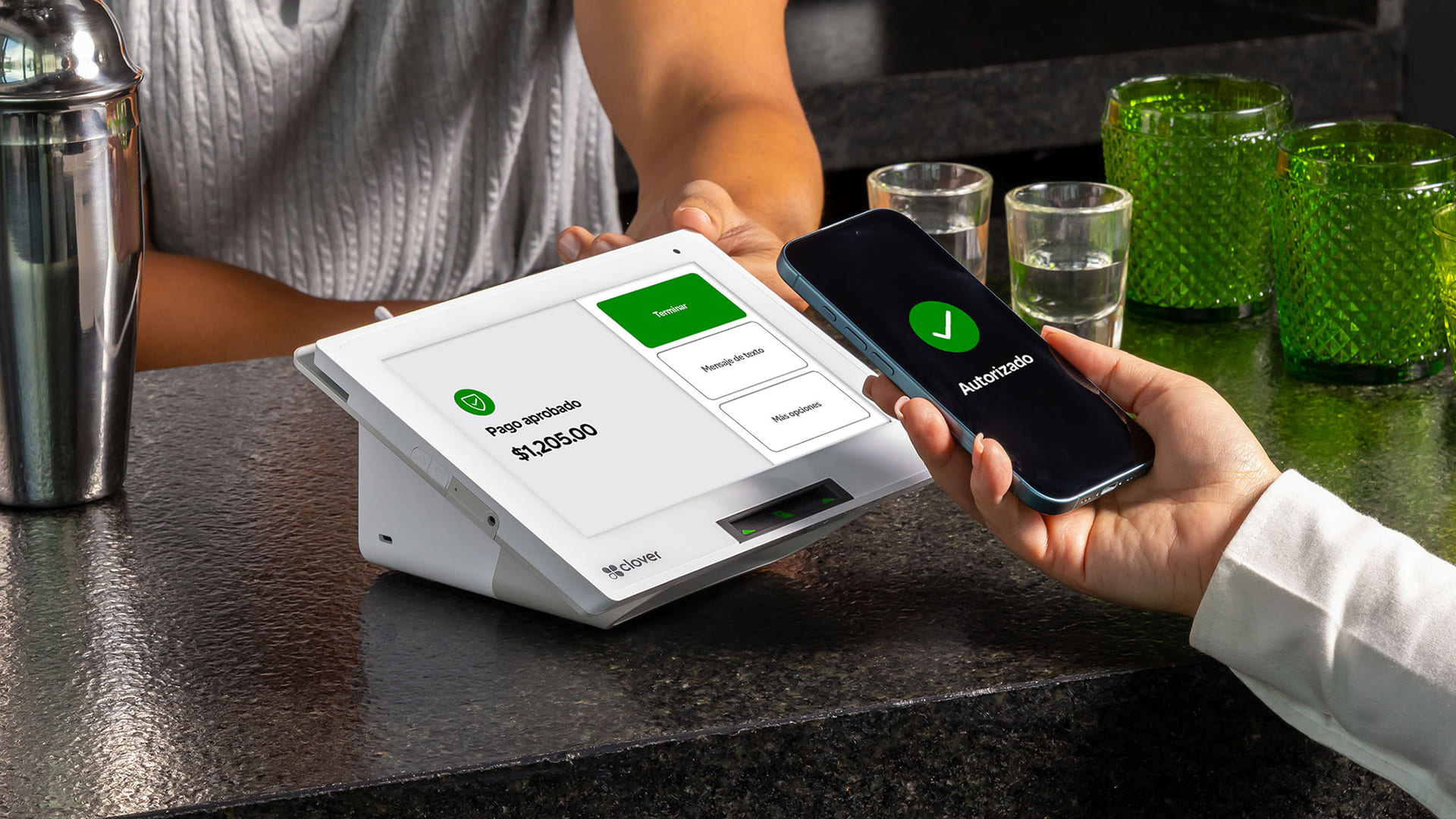 Una persona utiliza un teléfono inteligente para realizar un pago sin contacto en un terminal digital moderno situado en un mostrador. La pantalla muestra la confirmación del pago.